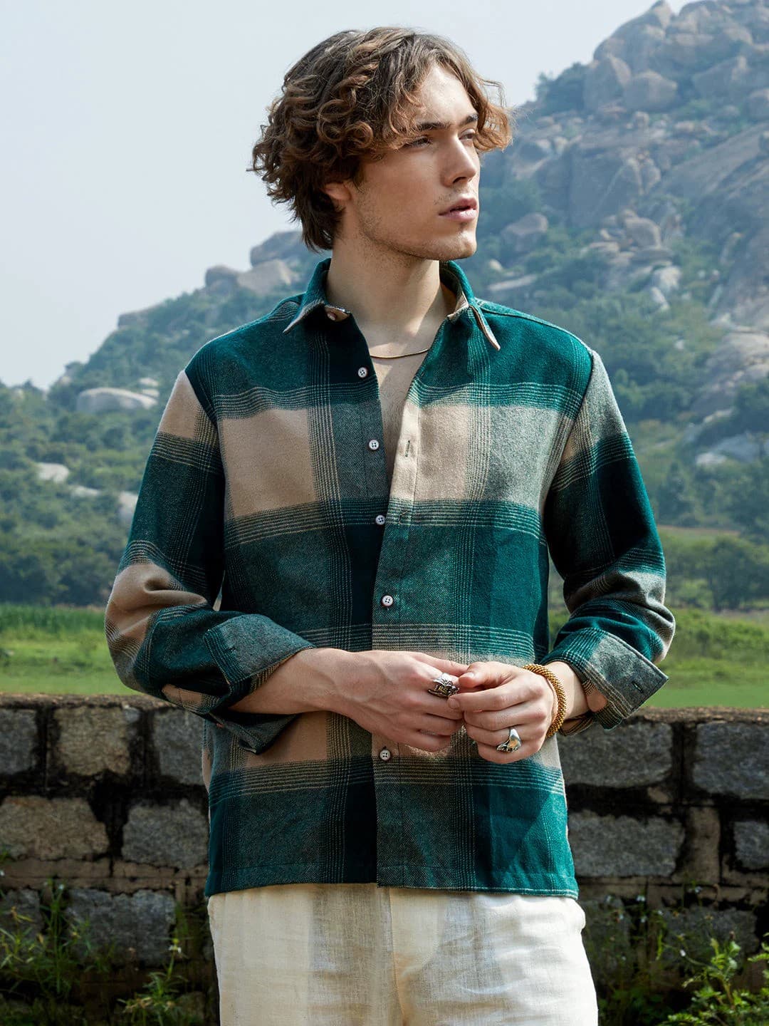 A man in a green and tan plaid overshirt, outdoors with hills behind him
